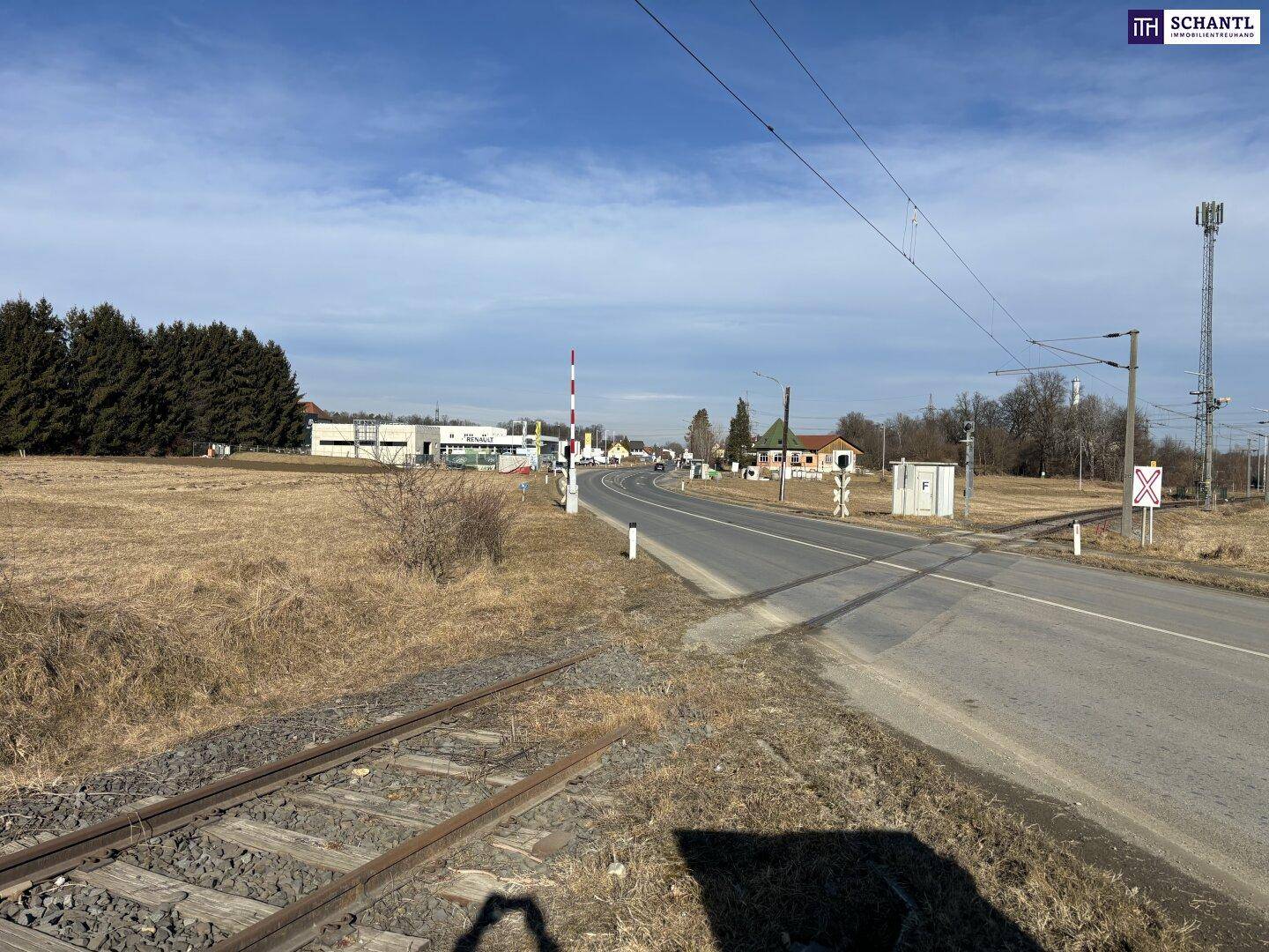 Trockenes Feld neben einer Landstraße und Bahngleisen, mit Bäumen und Gebäuden in der Ferne.