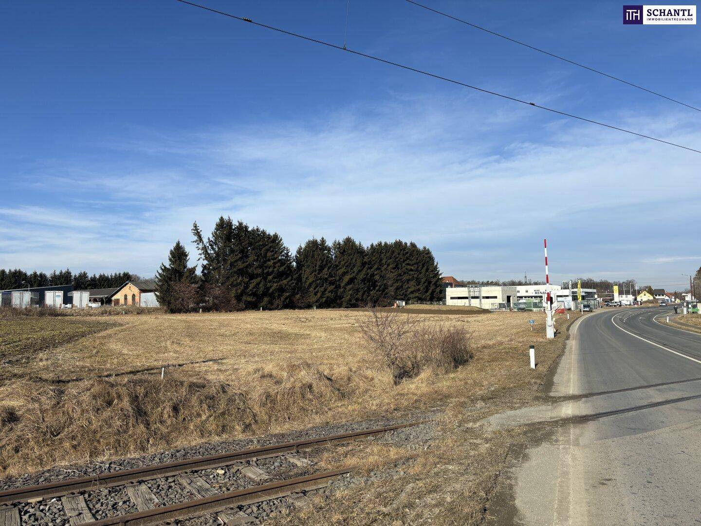 Weitläufiges trockenes Feld grenzt an eine Straße und Bahngleise, mit Bäumen und Gebäuden am Horizont.