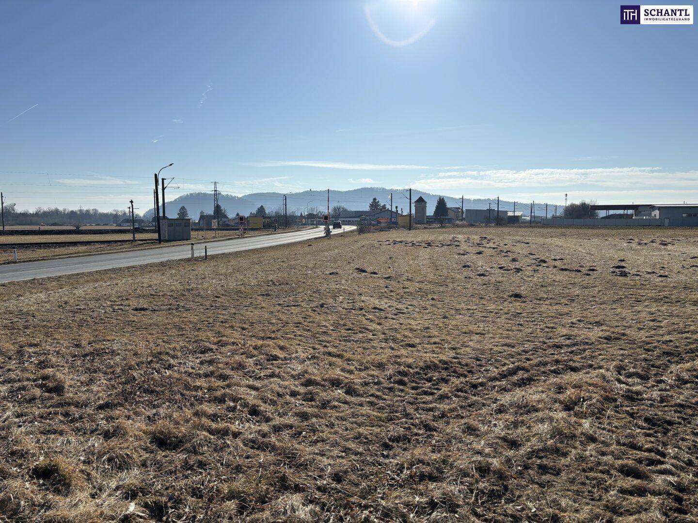 Weites, trockenes Feld neben einer Straße mit einer Bergkette in der Ferne unter strahlend blauem Himmel.