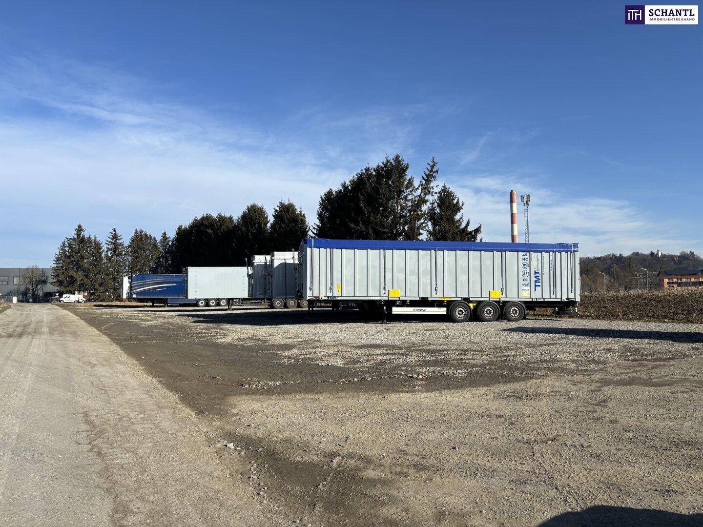 Großer Schotterparkplatz mit mehreren Lastwagenanhängern, umgeben von Bäumen und industriellen Strukturen.