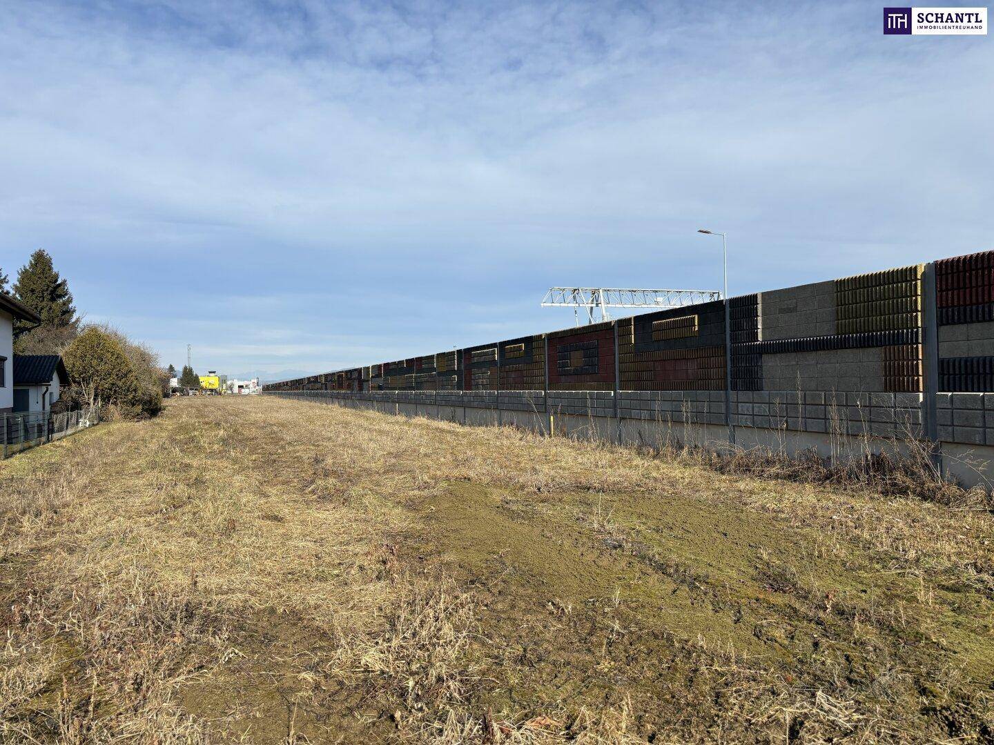 Weitläufiges, unbebautes Grundstück angrenzend an eine lange Lärmschutzwand mit Häusern in der Ferne.