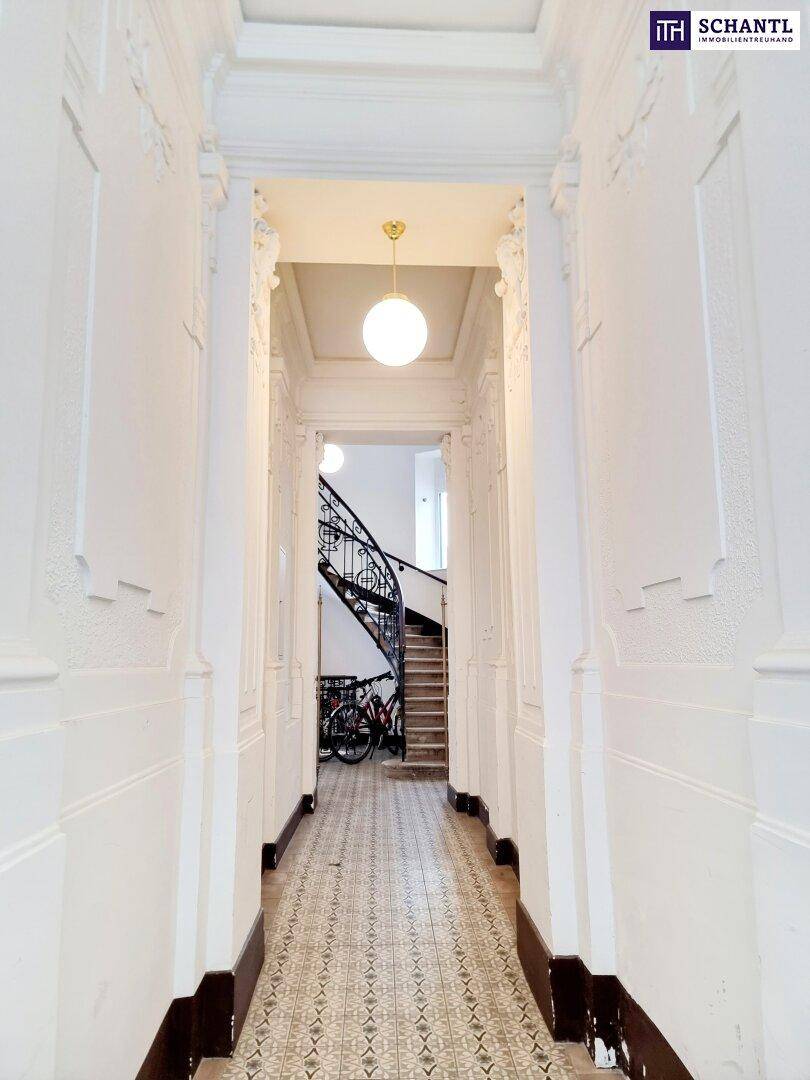 Langer, heller Flur mit gemusterten Fliesen und Stuckverzierungen an den Wänden, führt zu einer Treppe.