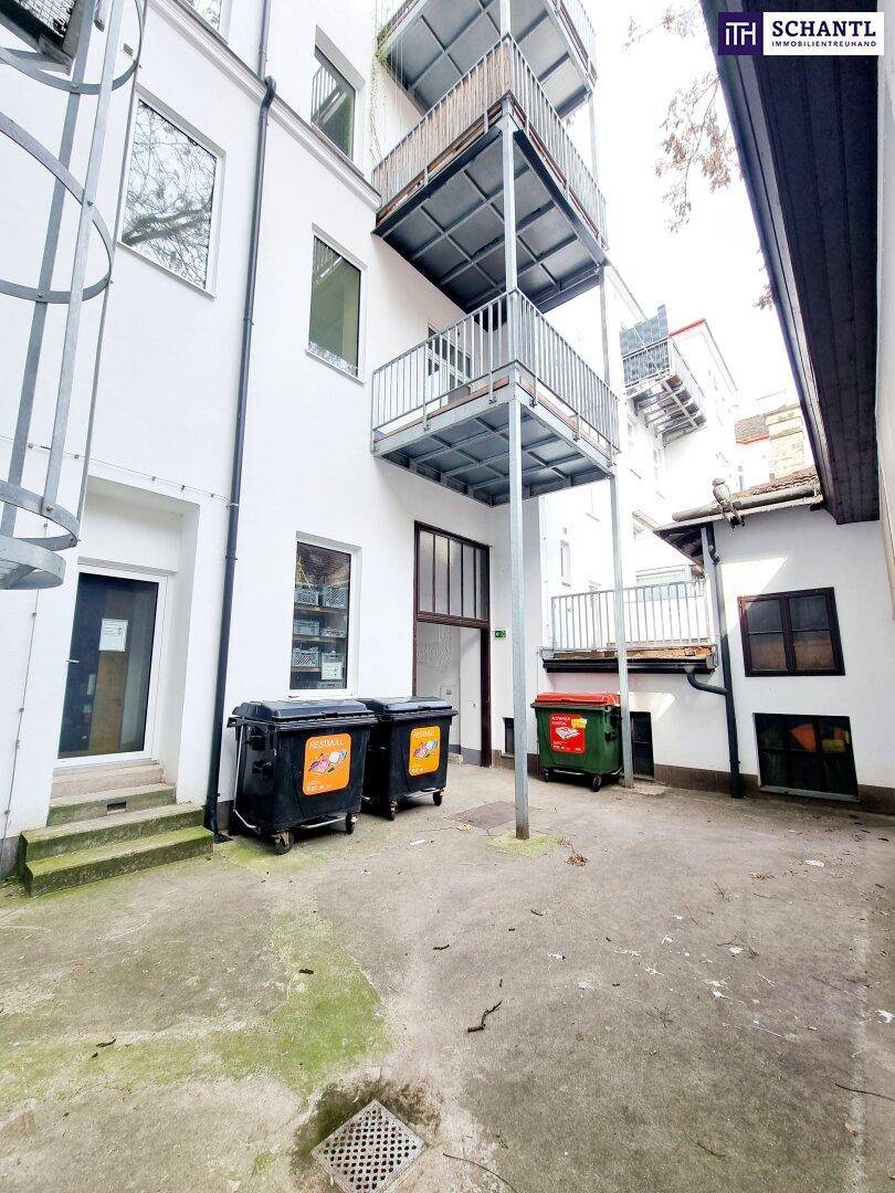 Hinterhofansicht mit Müllcontainern und einem Balkon im oberen Bereich, umgeben von Gebäudewänden.