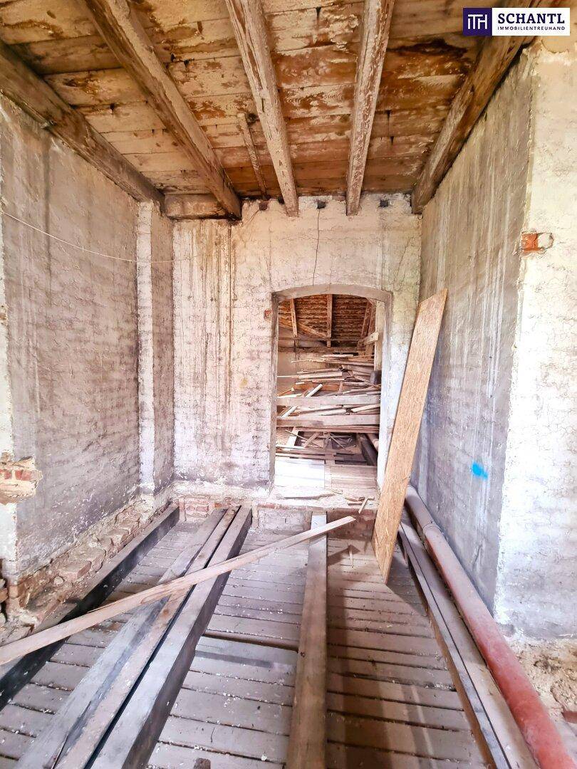 Langer Rohbau-Raum mit sichtbaren Holzbalken und einem Durchgang zum Dachboden.