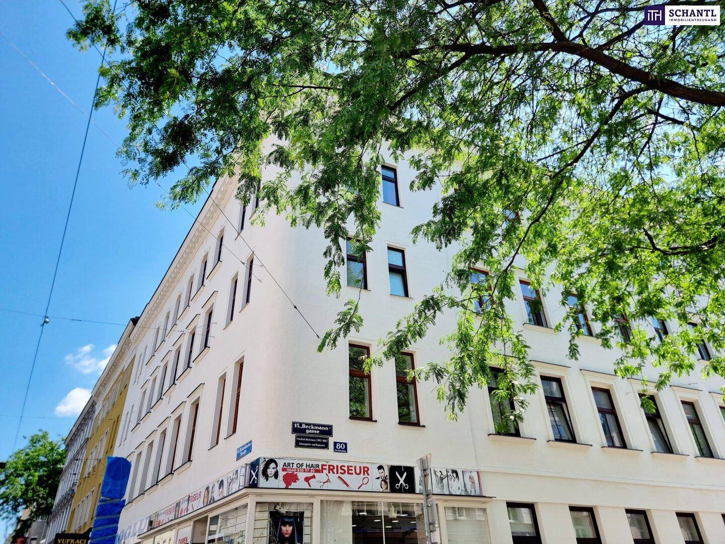 Historisches Eckgebäude mit Geschäften im Erdgeschoss und grünen Bäumen davor.