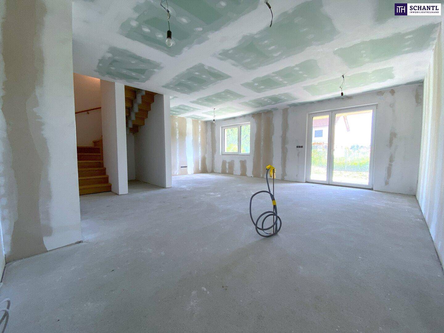 Unfertiger offener Wohnbereich mit Betonboden, Trockenbauwänden und Holztreppe.
