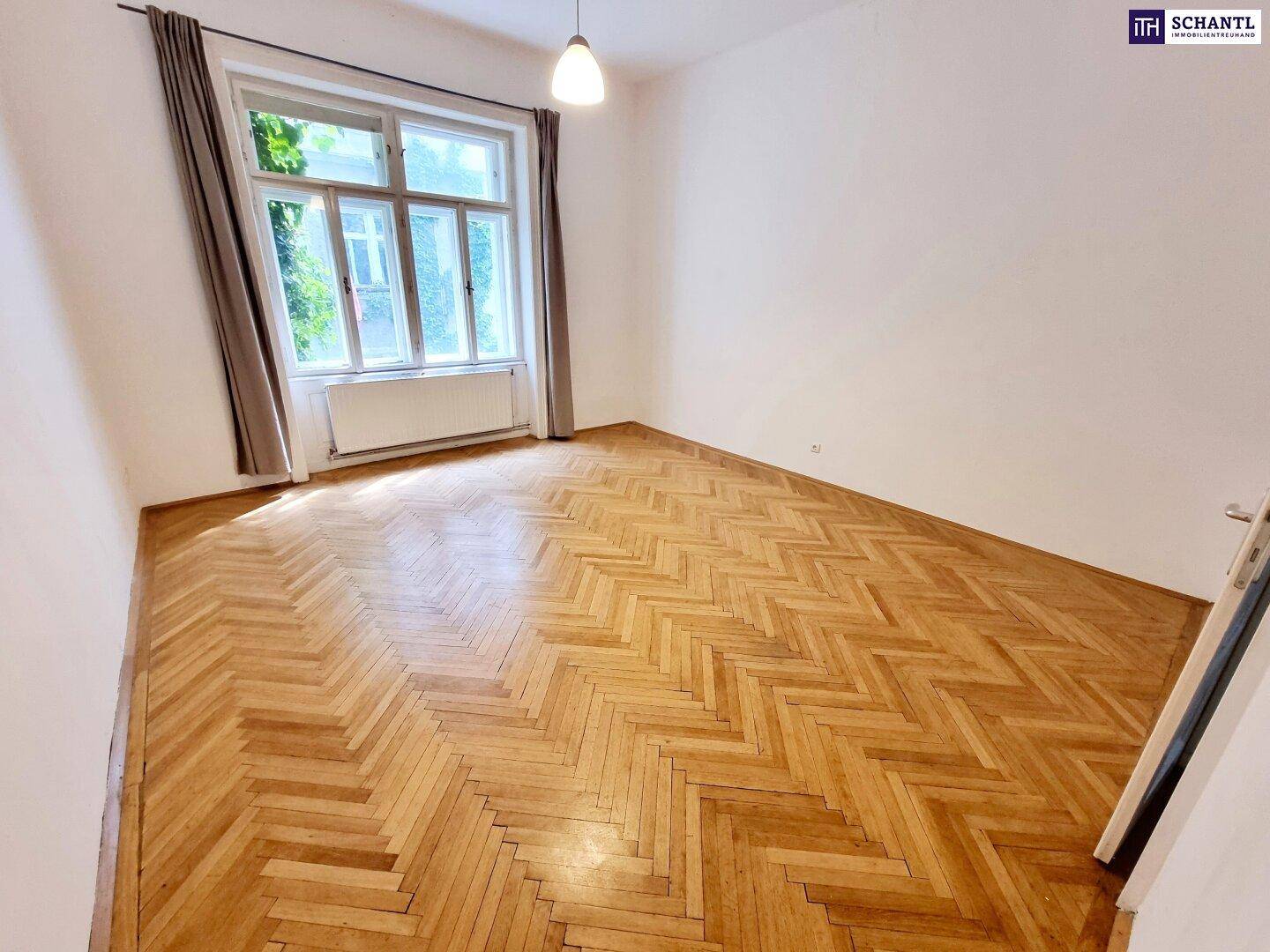 Lichtdurchflutetes Zimmer mit Fischgrätparkett, weißen Wänden und einem großen Fenster mit Vorhängen.
