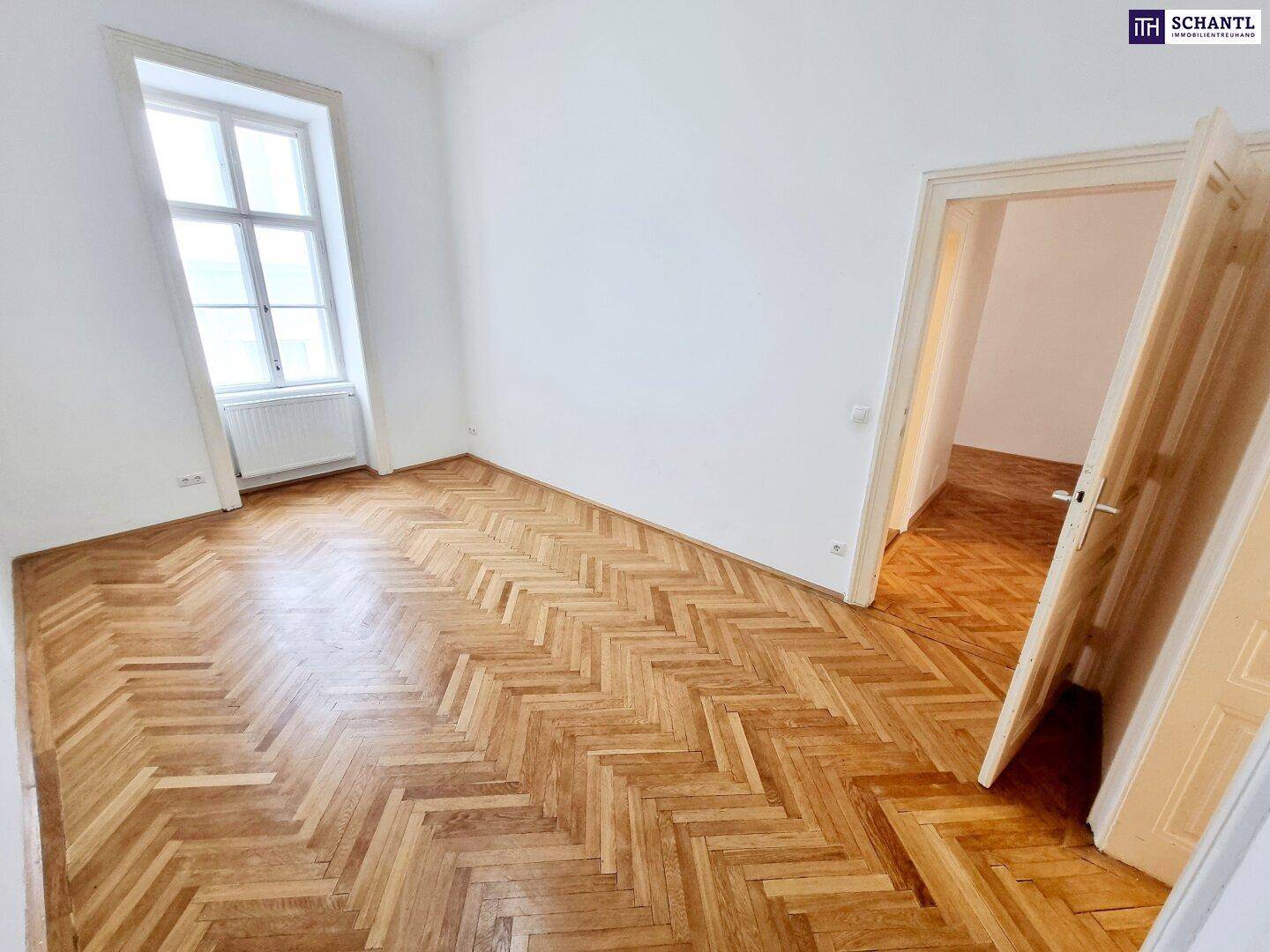 Geräumiges Zimmer mit klassischem Fischgrätparkett, hellen Wänden und Blick ins Nebenzimmer.