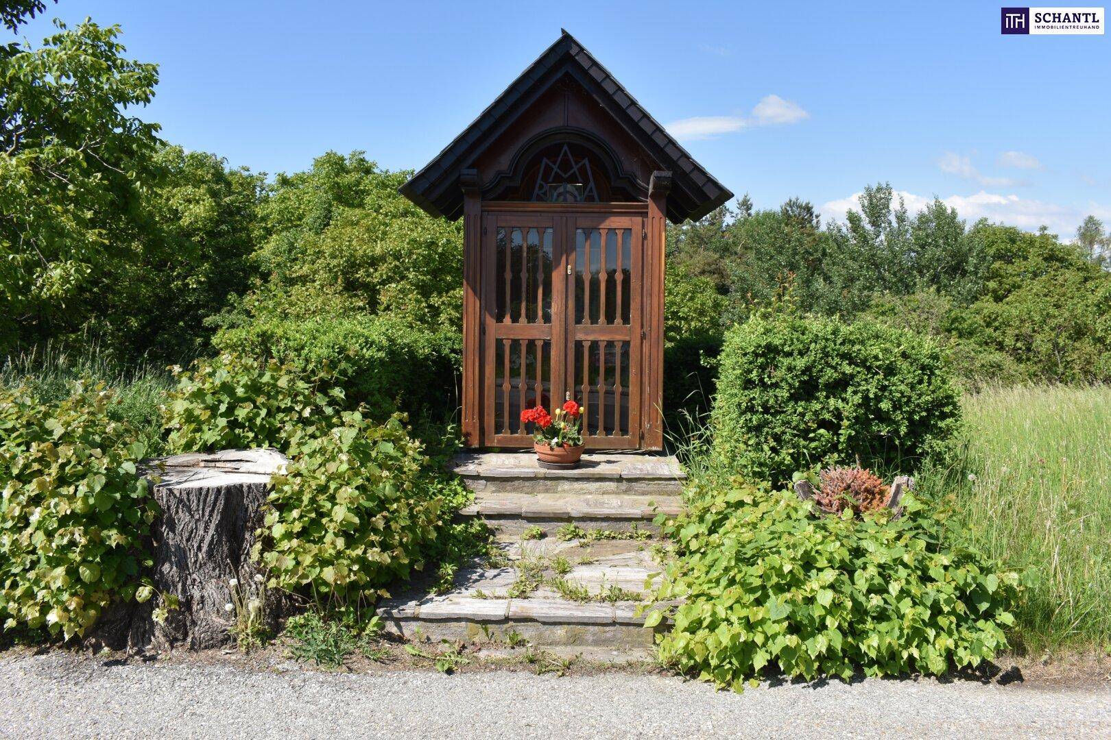 Kleiner hölzerner Schrein mit Blumen und üppiger grüner Vegetation in ländlicher Umgebung.