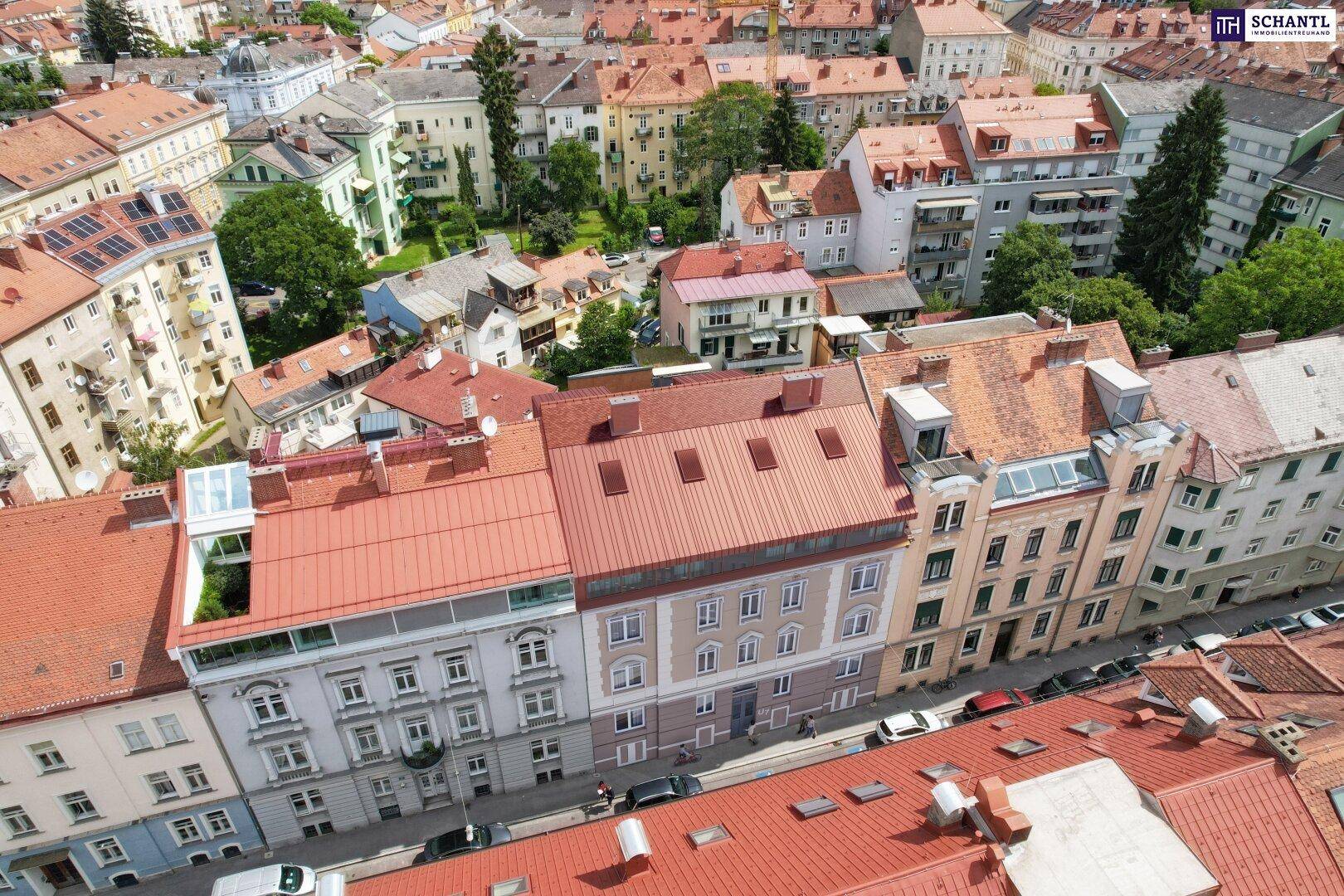 Panoramablick über eine dicht bebaute Stadtlandschaft mit zahlreichen Gebäuden und roten Dächern, umgeben von Grünflächen.