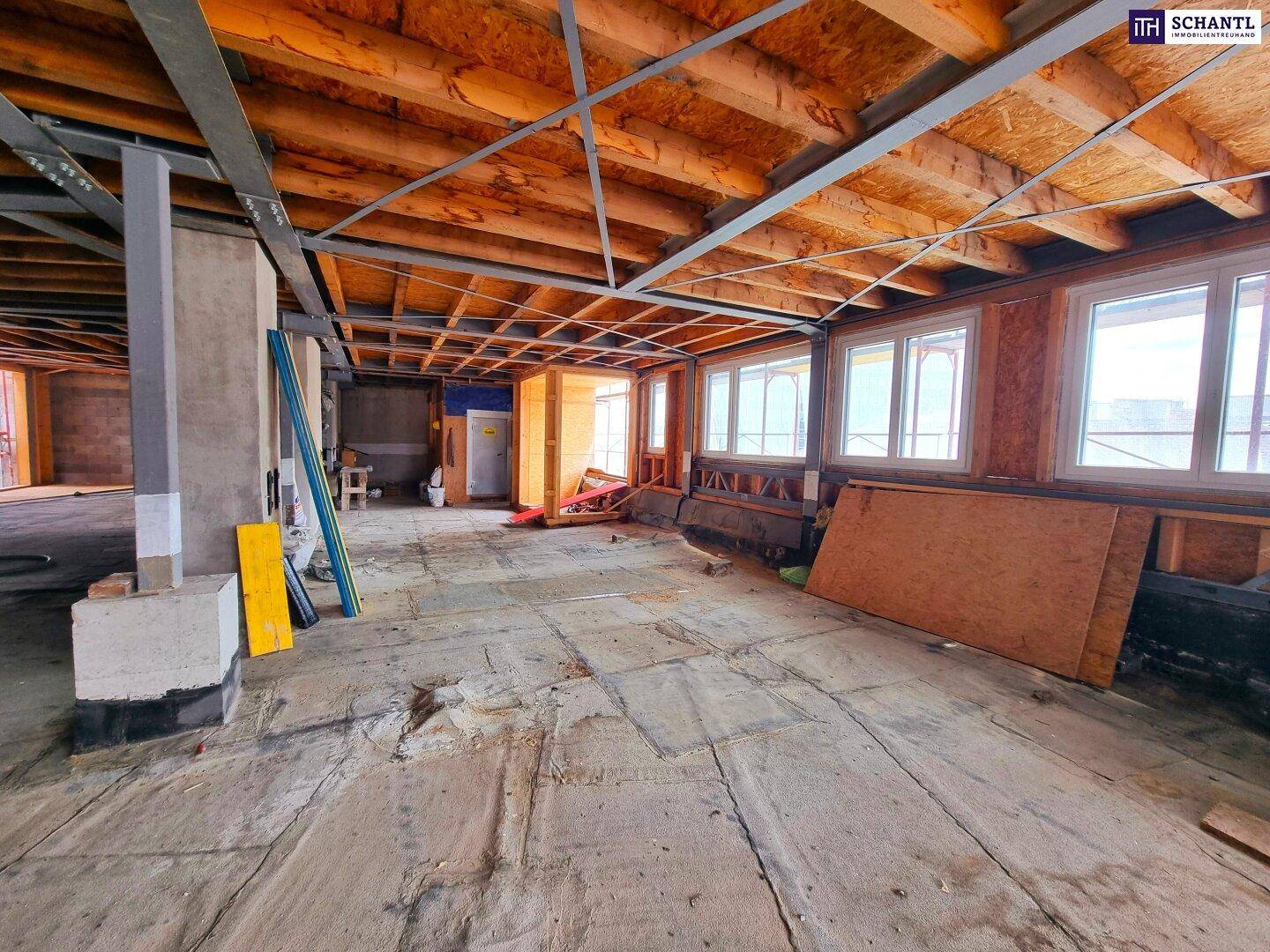 Großer Innenraum im Rohbau mit sichtbaren Holzdeckenbalken und vielen Fenstern, bereit für den Ausbau.