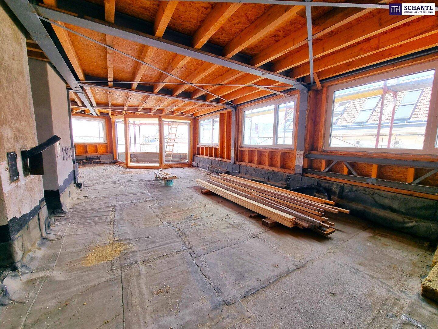 Geräumiger Rohbau im Dachgeschoss mit sichtbaren Holzbalken, großen Fensterfronten und Baumaterialien.
