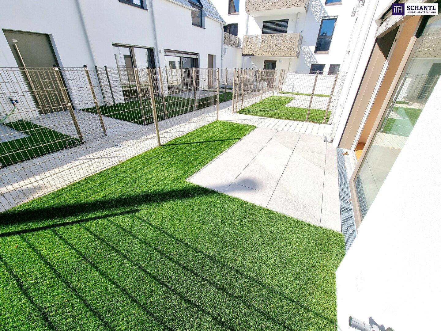 Neu angelegter Gartenbereich mit Kunstrasen und gepflasterter Terrasse, umgeben von modernen Gebäuden.