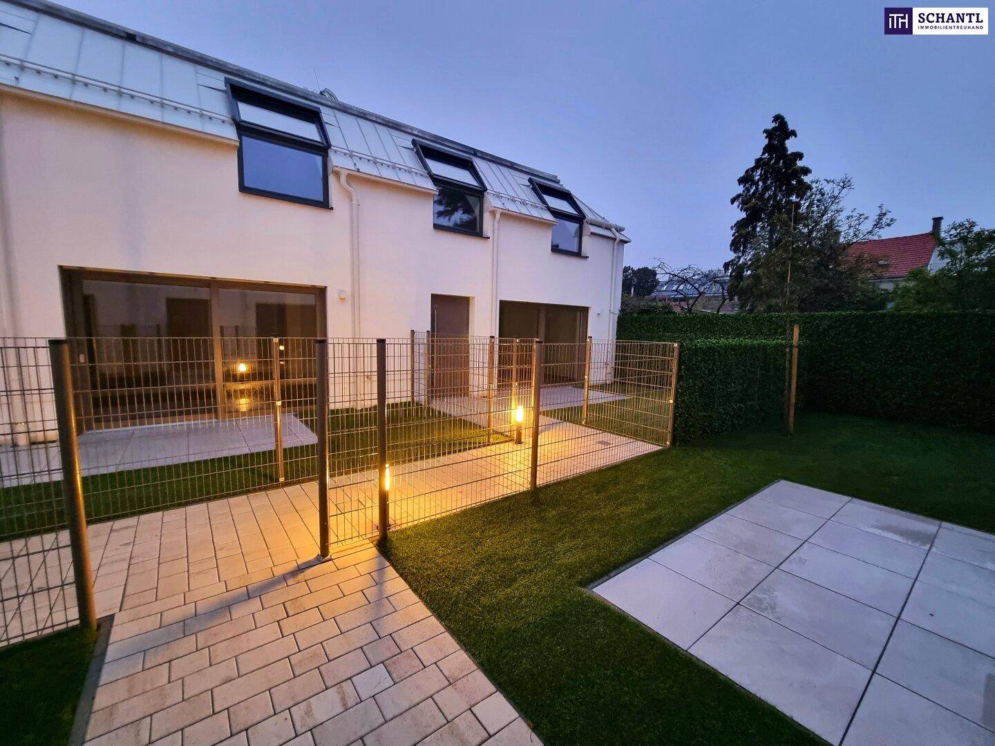 Gartenbereich mit Kunstrasen und gepflasterter Terrasse bei Abenddämmerung mit Beleuchtung.