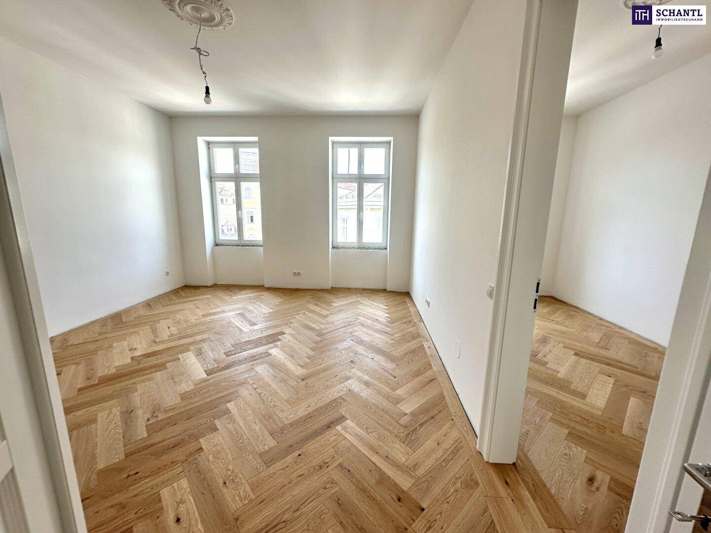 Großer, heller Raum mit Fischgrätparkett und zwei Fenstern, der viel natürliches Licht hereinlässt.