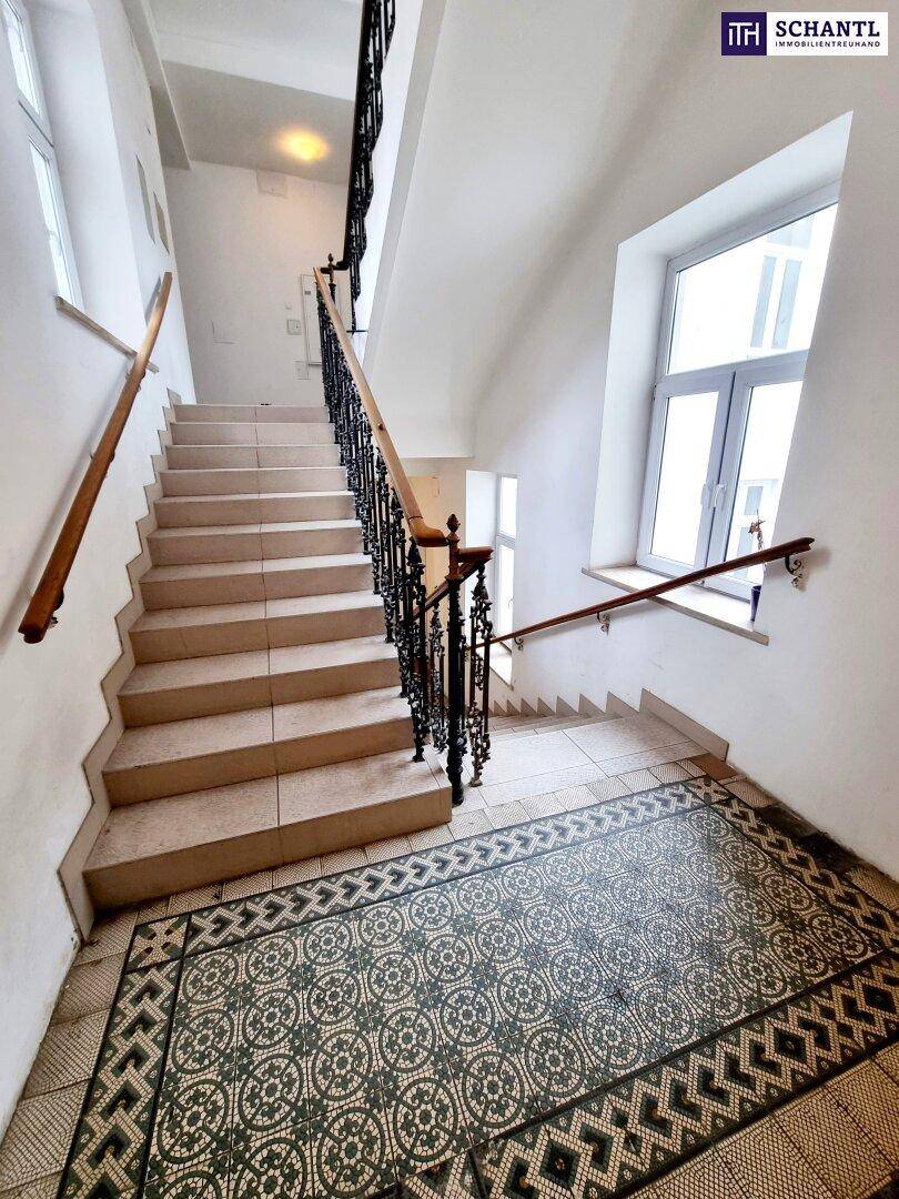 Helles Treppenhaus mit großen Fenstern, kunstvollem Geländer und gemusterten Fliesenböden.
