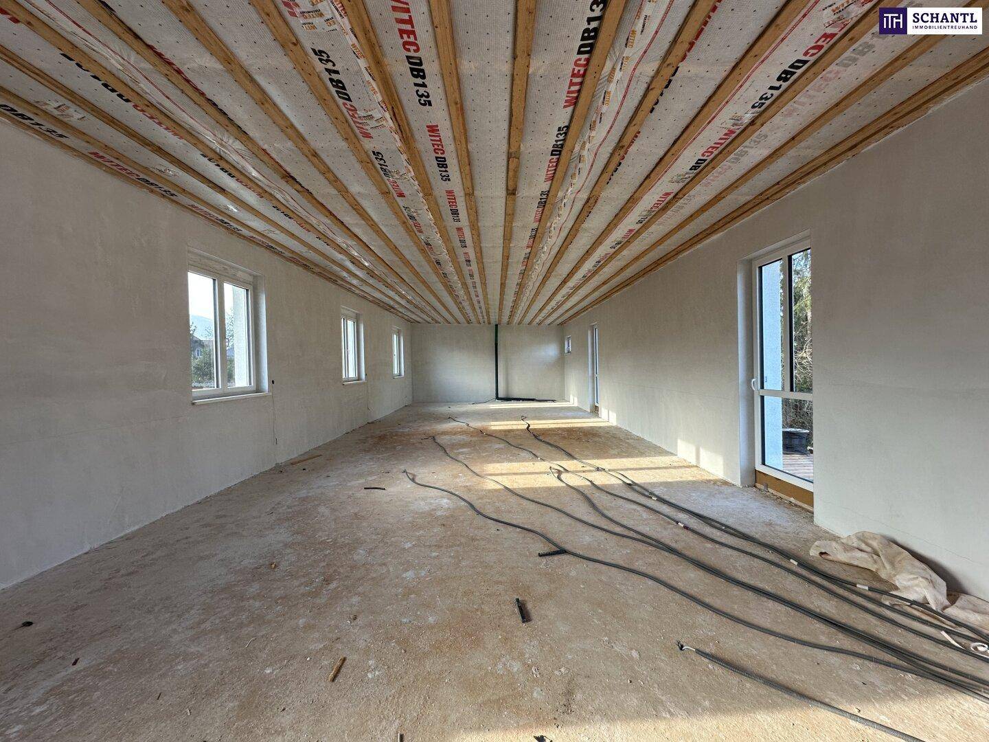 Geräumiger Innenraum im Rohbau mit sichtbarer Holzbalkendecke und unfertigem Betonboden.