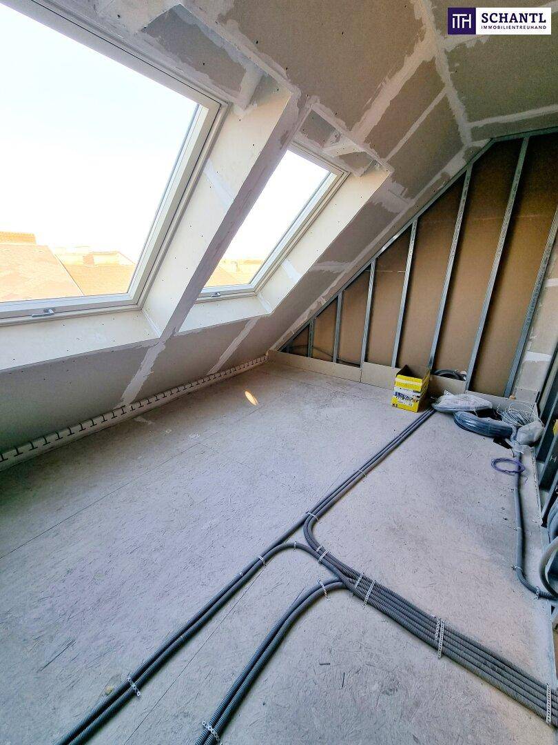 Dachgeschossraum im Rohbau mit schrägen Wänden, Dachfenstern und sichtbaren Leitungen.