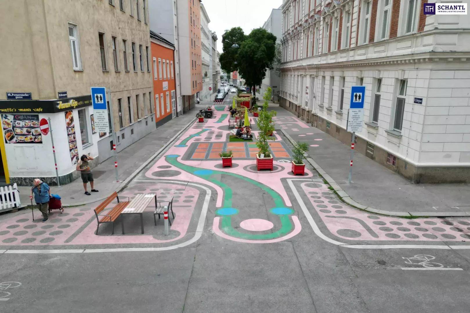 Belebte Fußgängerzone mit farbenfroher Bodengestaltung, Sitzgelegenheiten und Cafés.