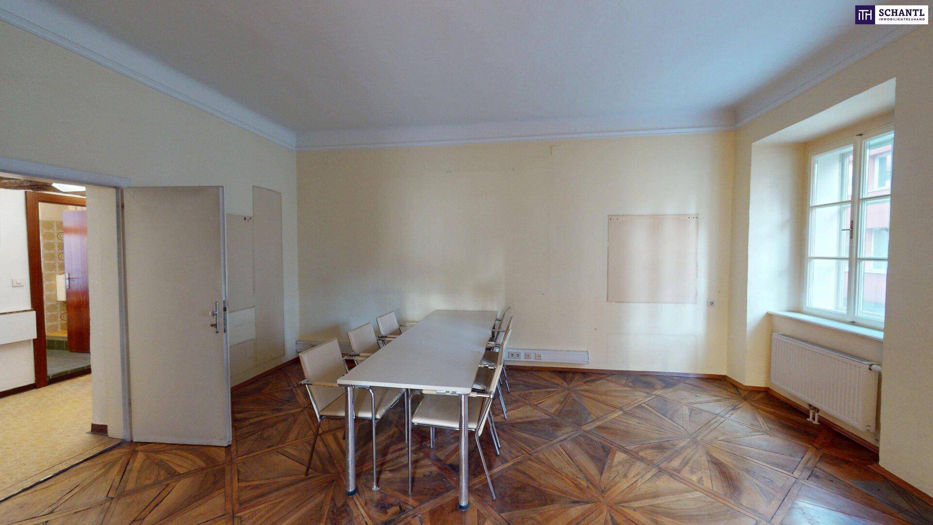 Besprechungsraum mit gemustertem Holzboden, großem Tisch und Stühlen, hell durch Fenster.