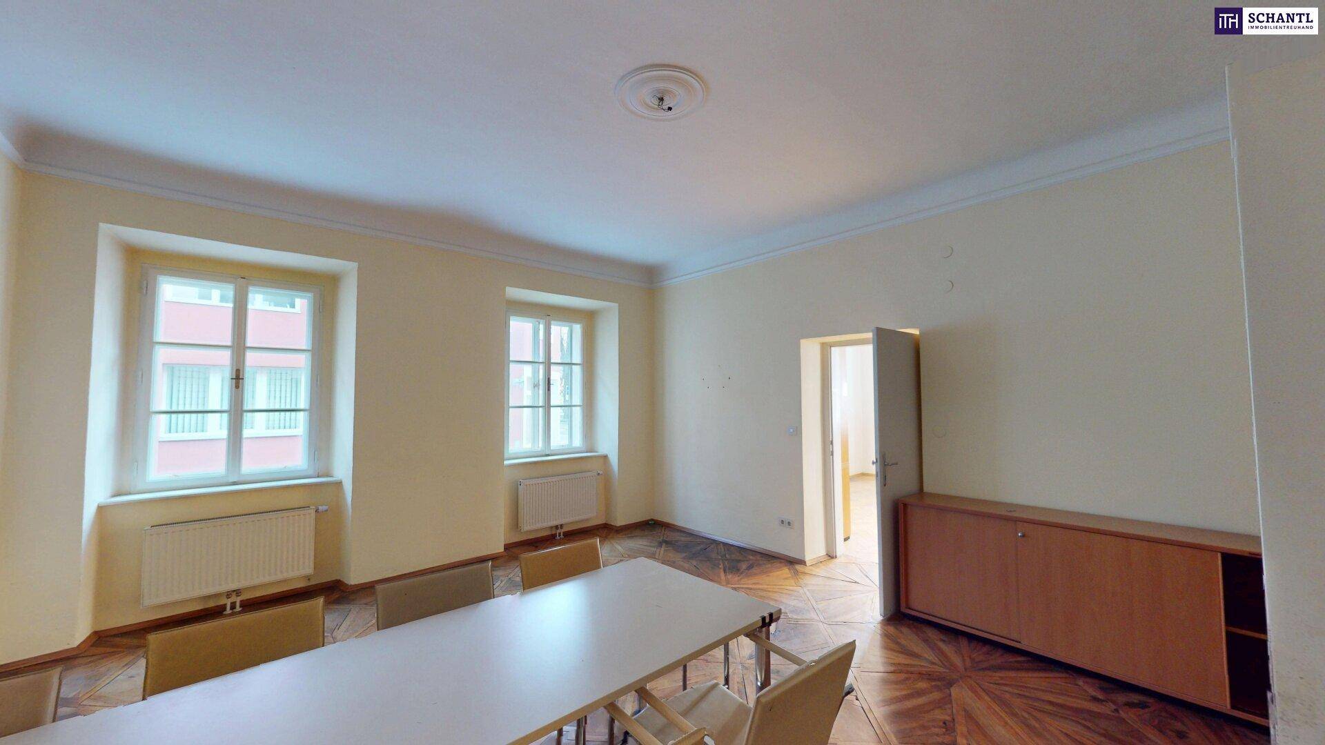 Besprechungsraum mit Holzboden, großem Tisch und Schränken, hell durch zwei Fenster.