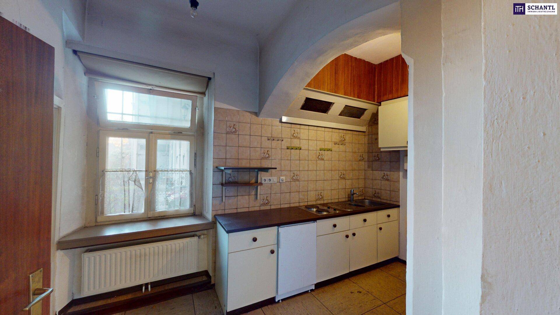 Ältere Küche mit Fliesenboden, Einbauschränken und Gasherd, hell durch ein Fenster.