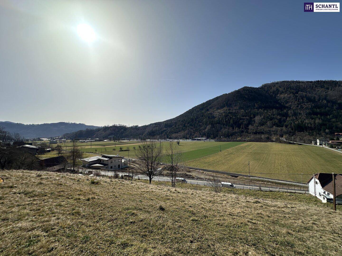 Sonnige Landschaft mit weiten Feldern, einer Straße und bewaldeten Bergen im Hintergrund.