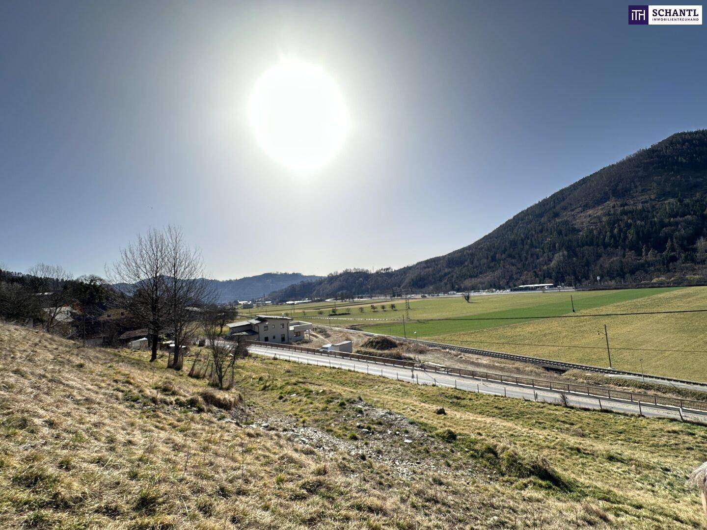 Weitläufige Landschaft mit grünen Feldern, einer Straße und bewaldeten Bergen unter strahlendem Sonnenschein.