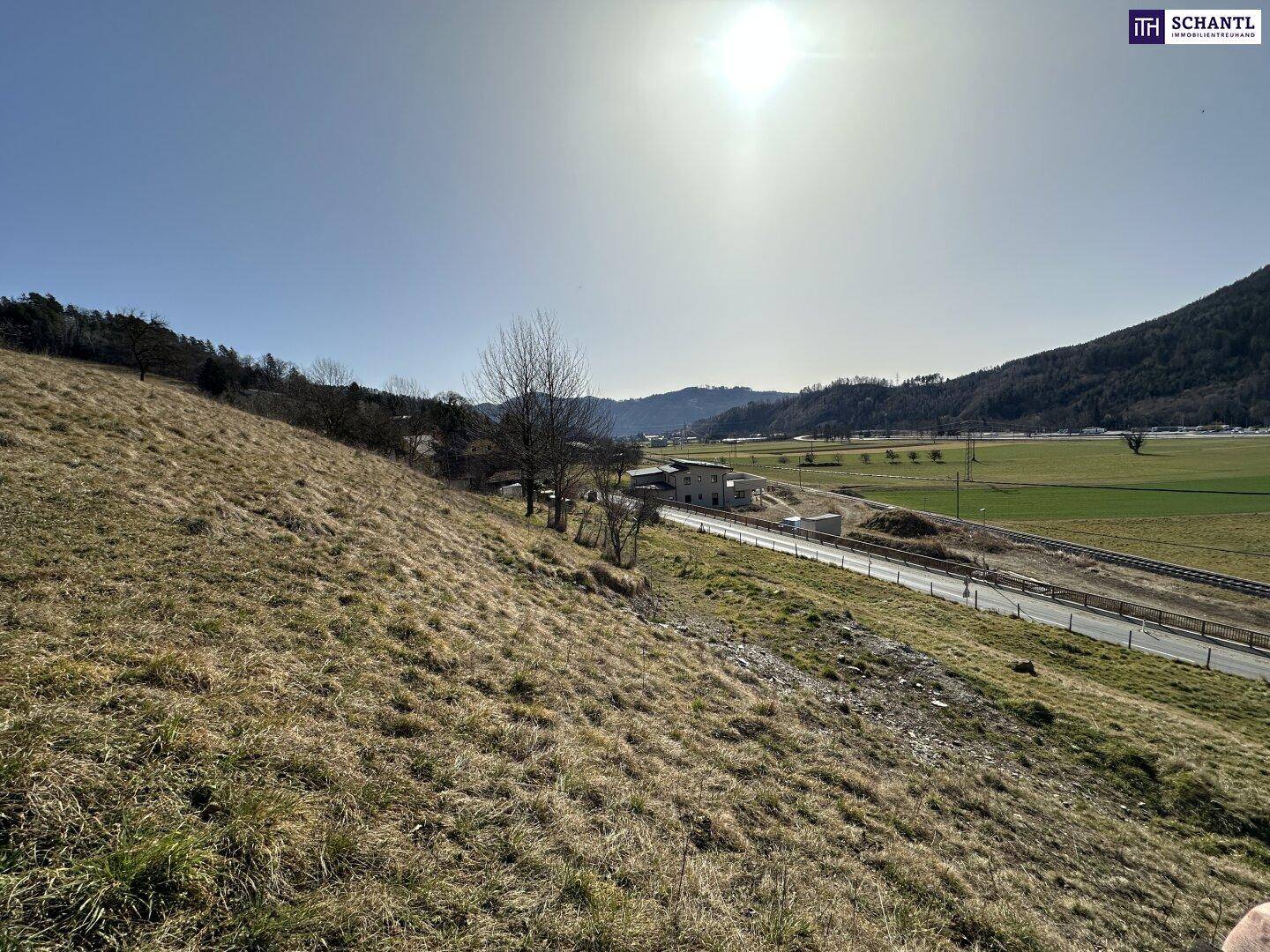 Sonnige Aussicht über eine weite Landschaft mit Feldern, einer Straße und Bergen am Horizont.