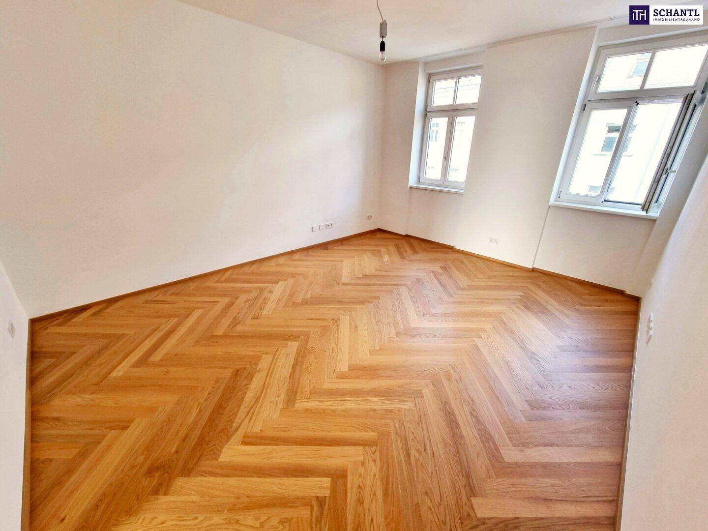 Leeres, helles Zimmer mit Fischgrätparkettboden und zwei großen Fenstern.