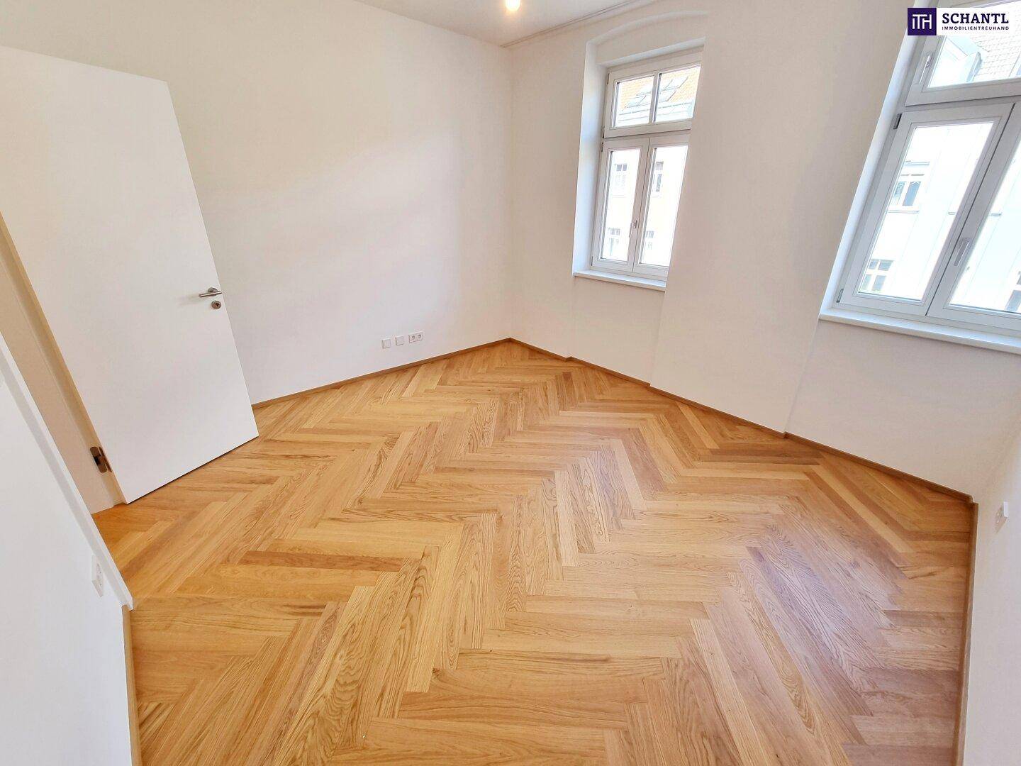 Helles Zimmer mit Fischgrätparkettboden und zwei großen Fenstern.