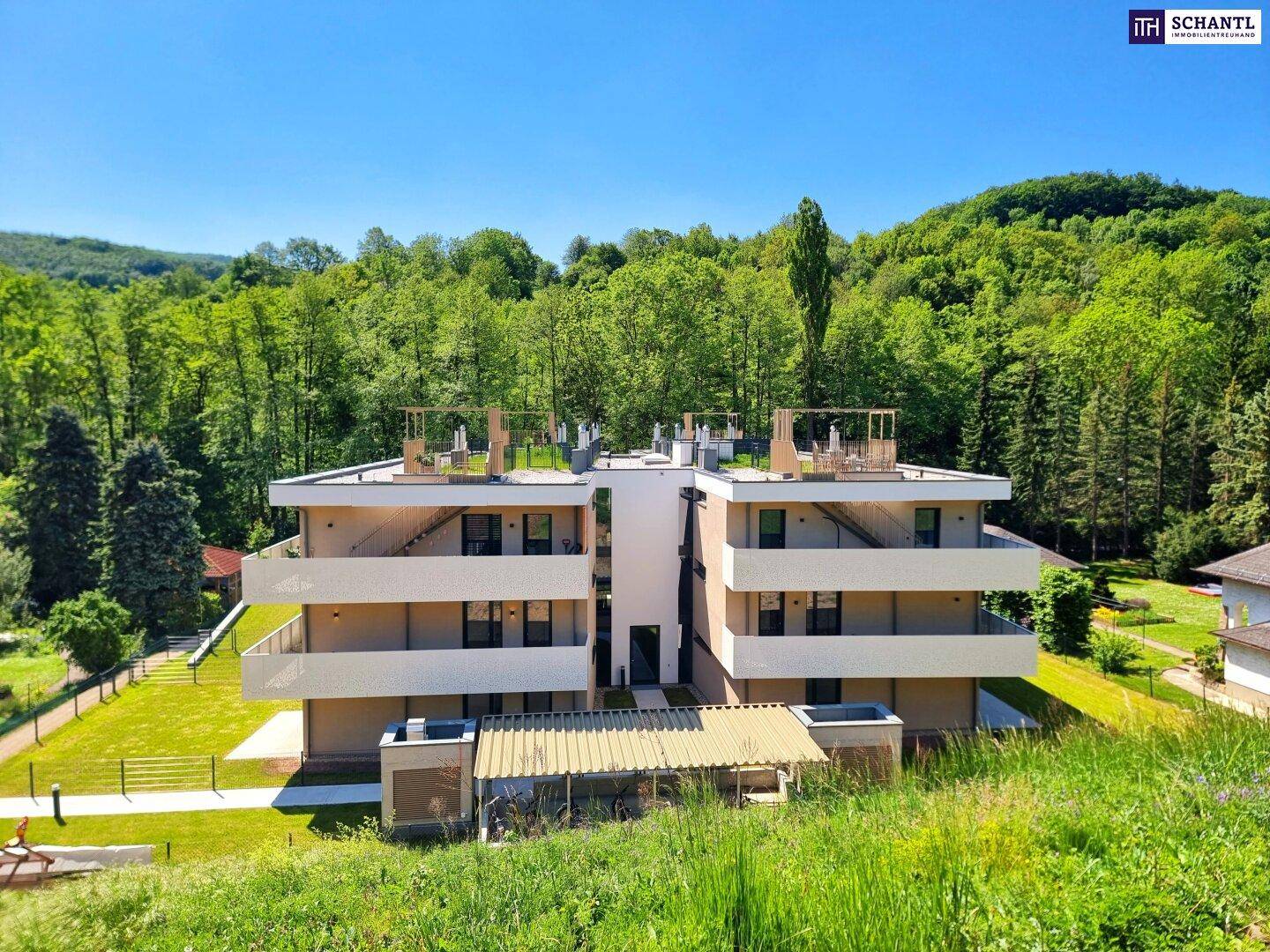 Modernes Mehrfamilienhaus in grüner Umgebung mit Balkonen und Dachterrassen unter blauem Himmel.