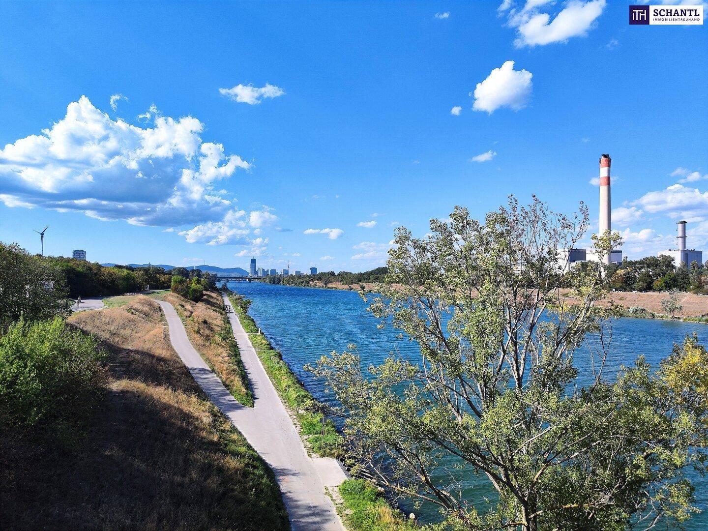 Flusslandschaft mit Uferweg, umgeben von Bäumen und Blick auf Industrieanlagen in der Ferne.