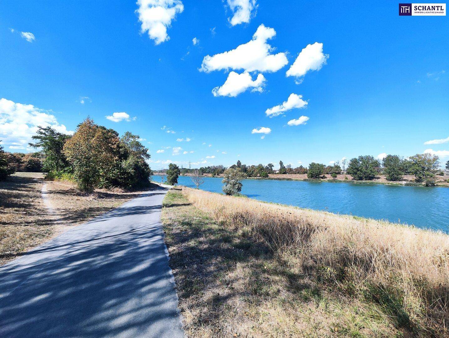 Uferweg entlang eines ruhigen Flusses, gesäumt von Bäumen und trockenem Gras unter blauem Himmel.