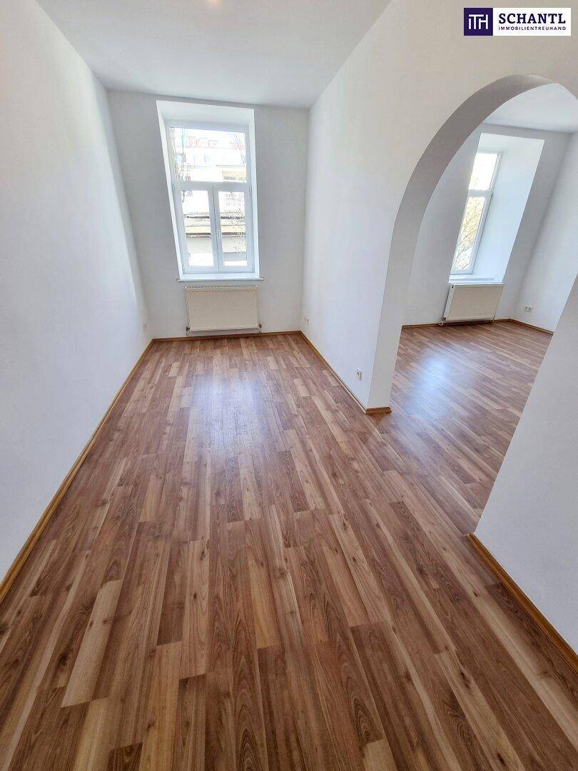 Geräumiges Wohnzimmer mit Laminatboden, hohen Fenstern und einem Rundbogen zu einem weiteren Bereich.