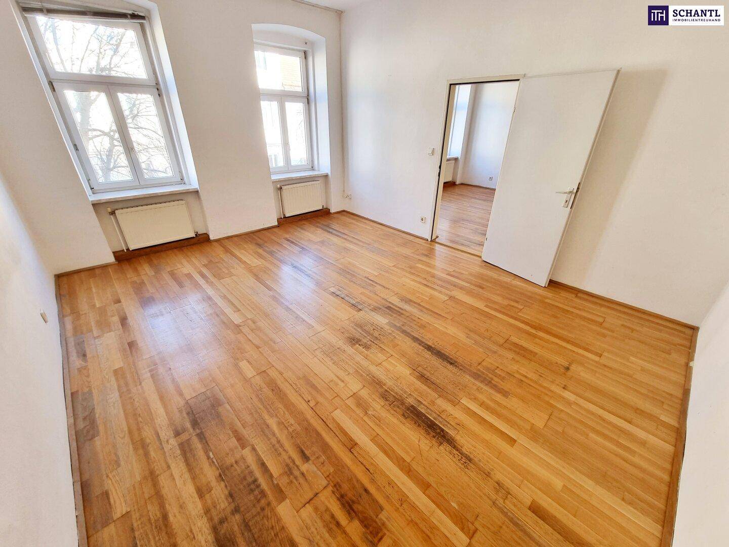 Heller Raum mit Holzboden, weißen Wänden und zwei Fenstern, die viel Licht spenden.