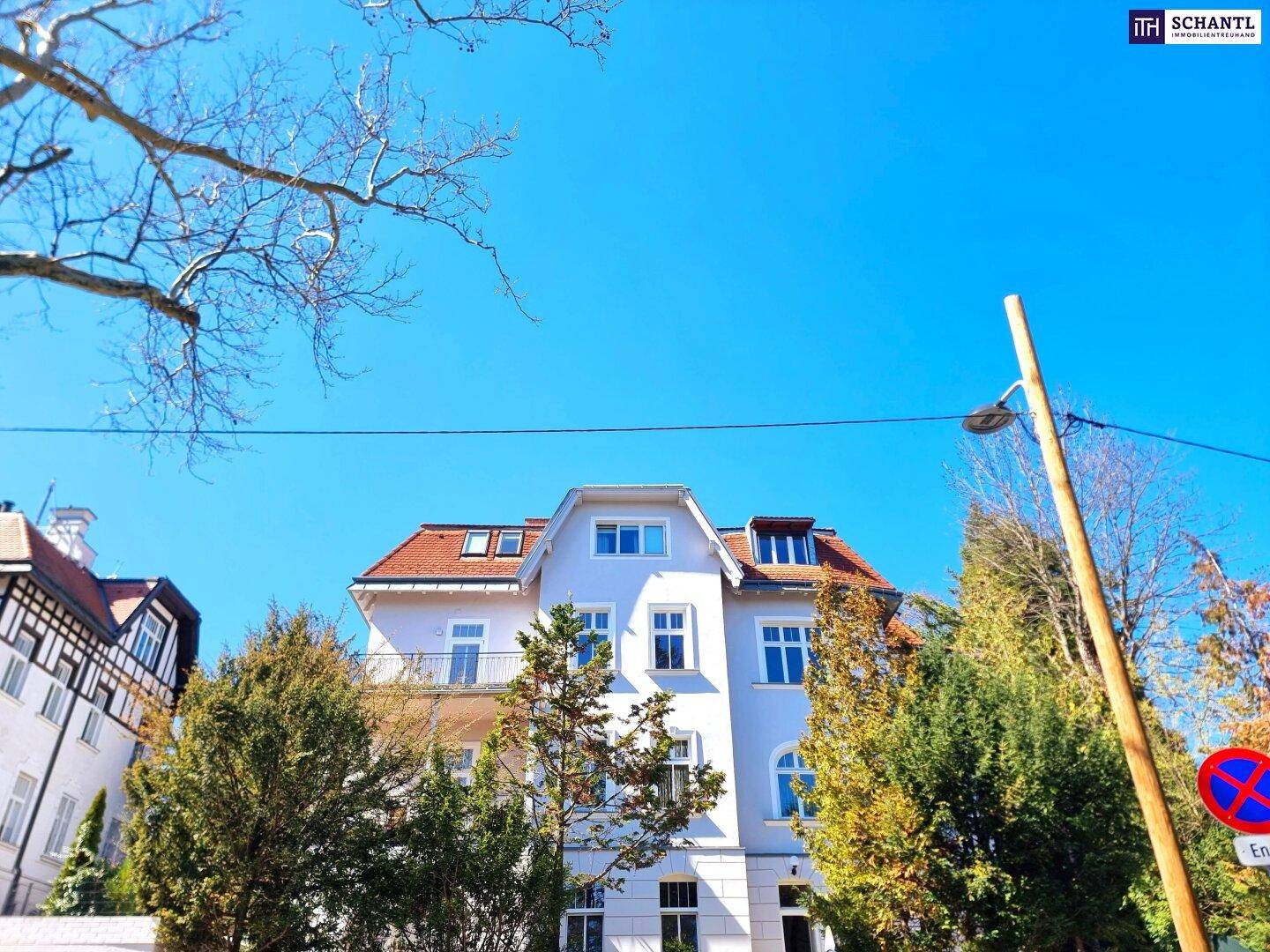 Historisches Gebäude mit klassischer Architektur und roten Dachziegeln, umgeben von Bäumen.