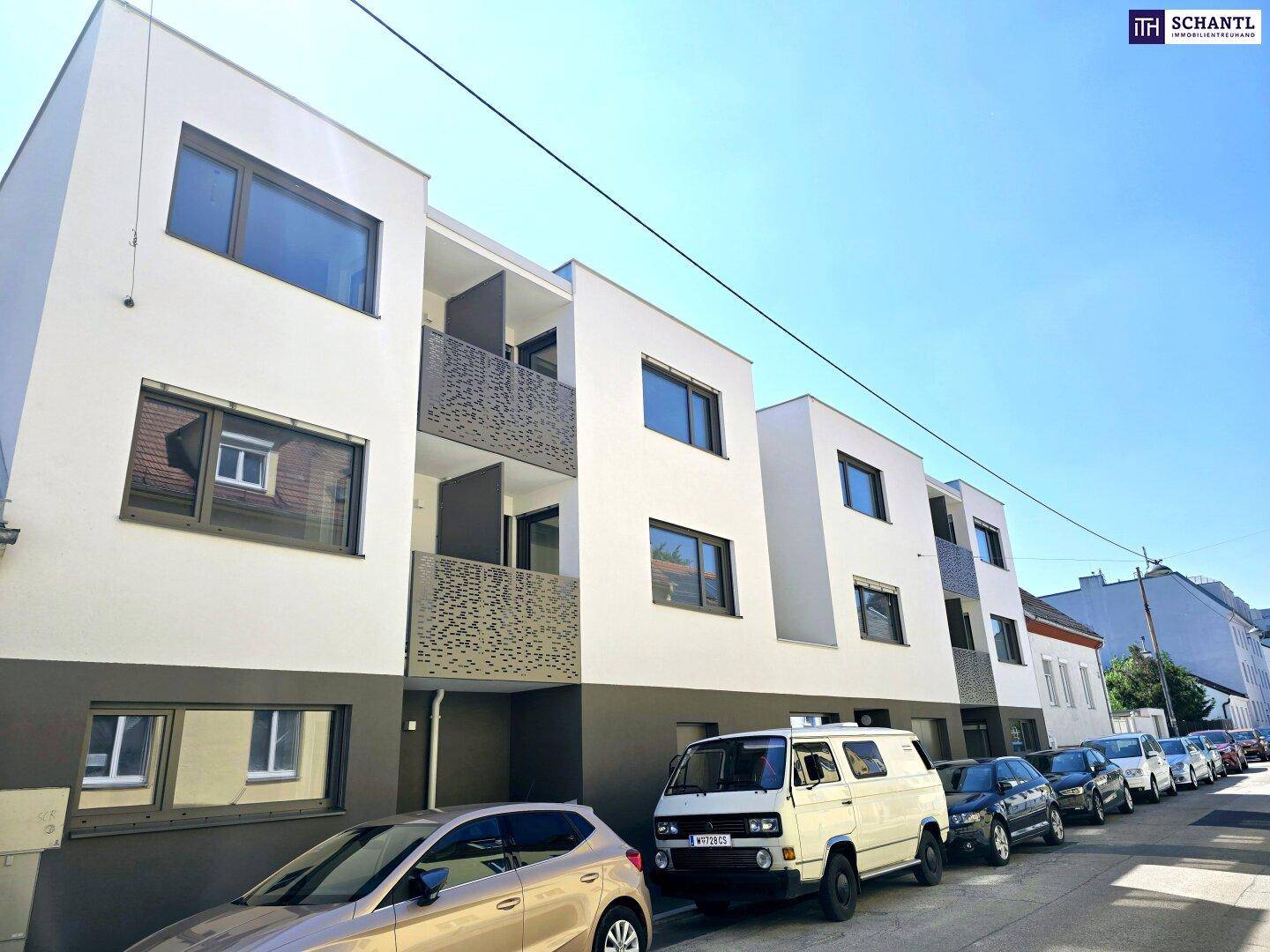 Ansicht einer modernen Wohnanlage mit weißer Fassade, Balkonen und parkenden Fahrzeugen auf der Straße.