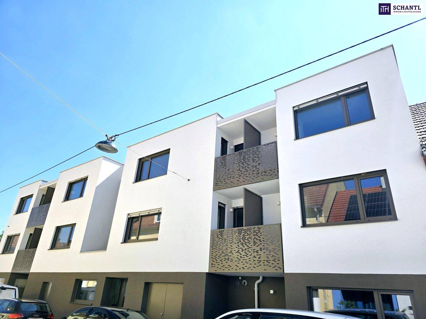 Moderne Wohnanlage mit weißer Fassade, Balkonen und parkenden Autos auf der Straße.