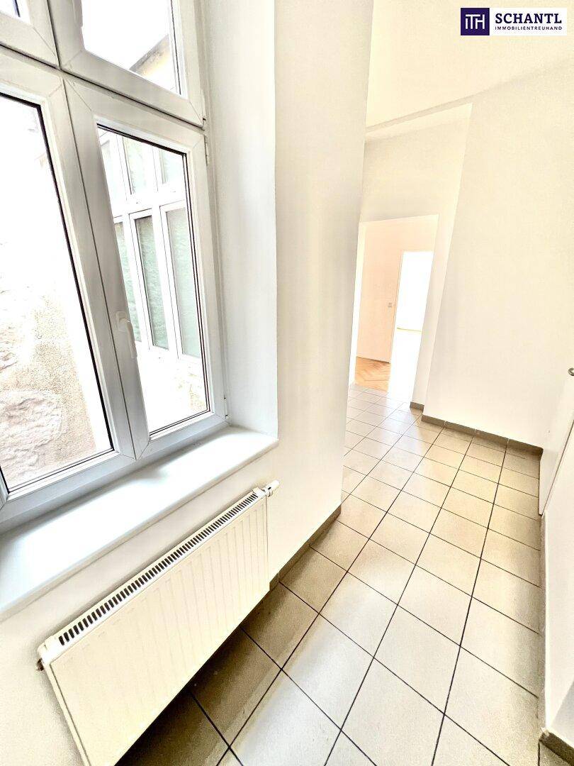 Heller Flur mit Fliesenboden, weiß gestrichenen Wänden und einem Fenster, das Tageslicht hereinlässt.