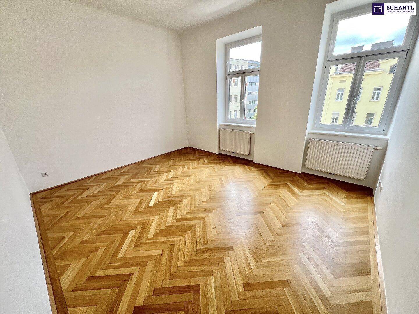Heller Raum mit klassischem Fischgrätparkett, hohen Fenstern und Heizkörpern unter den Fenstern.
