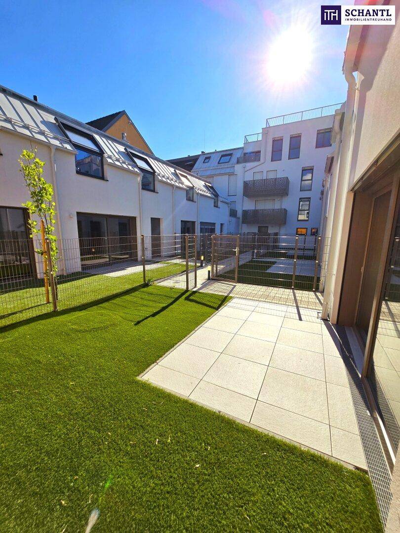 Sonnige Gartenfläche mit Kunstrasen und gepflasterter Terrasse in einer Wohnanlage.