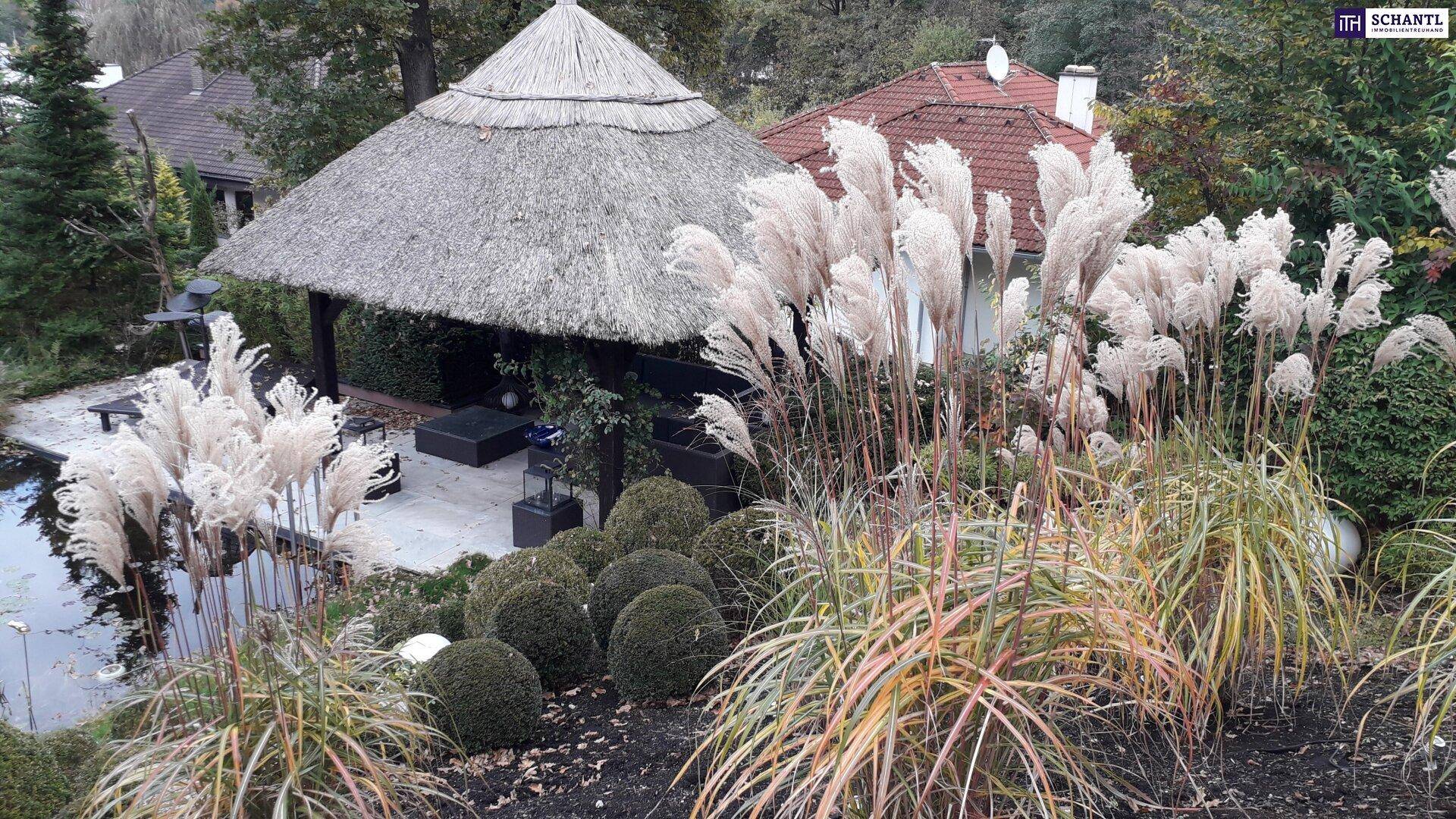 Gepflegter Garten mit Ziergräsern und einem traditionellen Strohdach-Pavillon, umgeben von Grün.