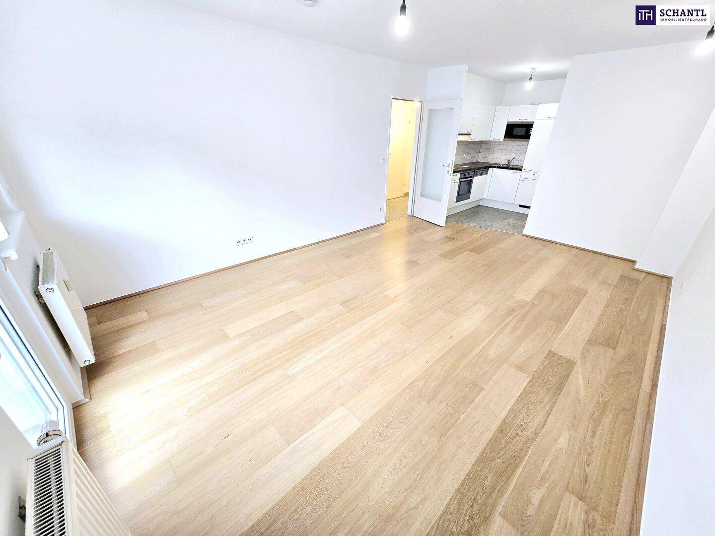 Geräumiges Wohnzimmer mit hellem Holzboden und offener Verbindung zu einer modernen Küchenzeile im Hintergrund.