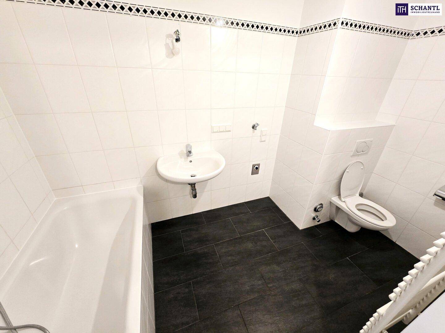 Badezimmer mit Badewanne, Waschbecken und Wand-WC, ausgestattet mit dunklen Bodenfliesen und weißer Wandverkleidung.