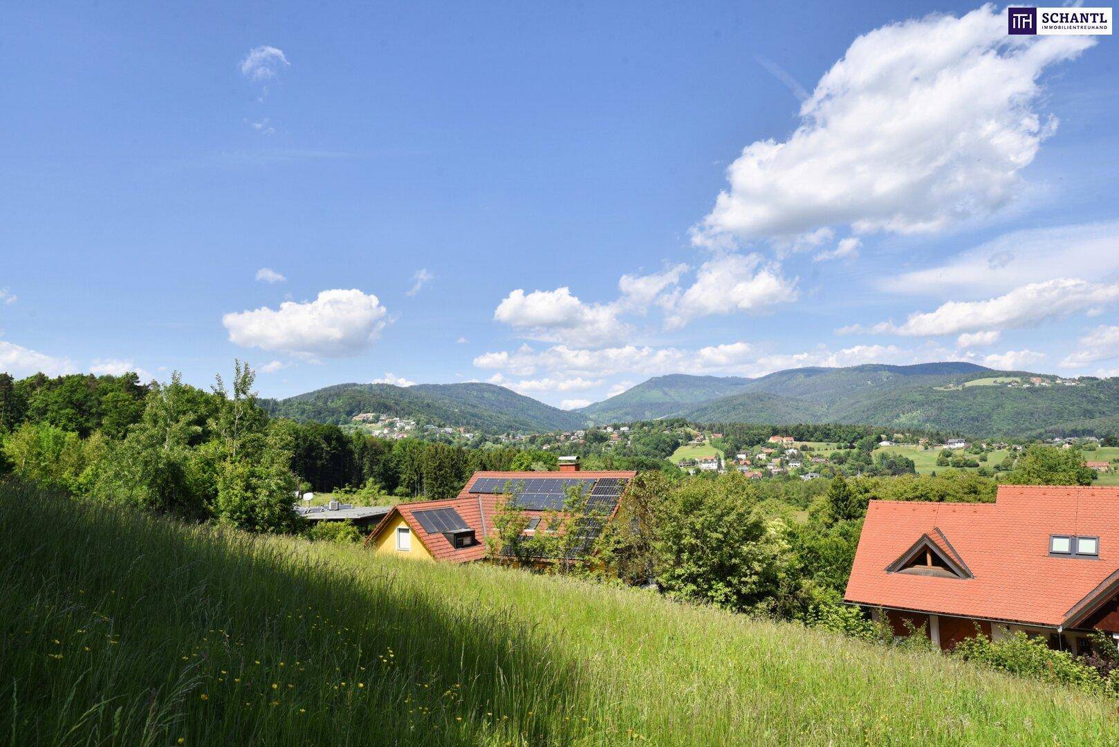Panoramablick über eine grüne Hügellandschaft mit Häusern und weiten Bergen unter blauem Himmel.