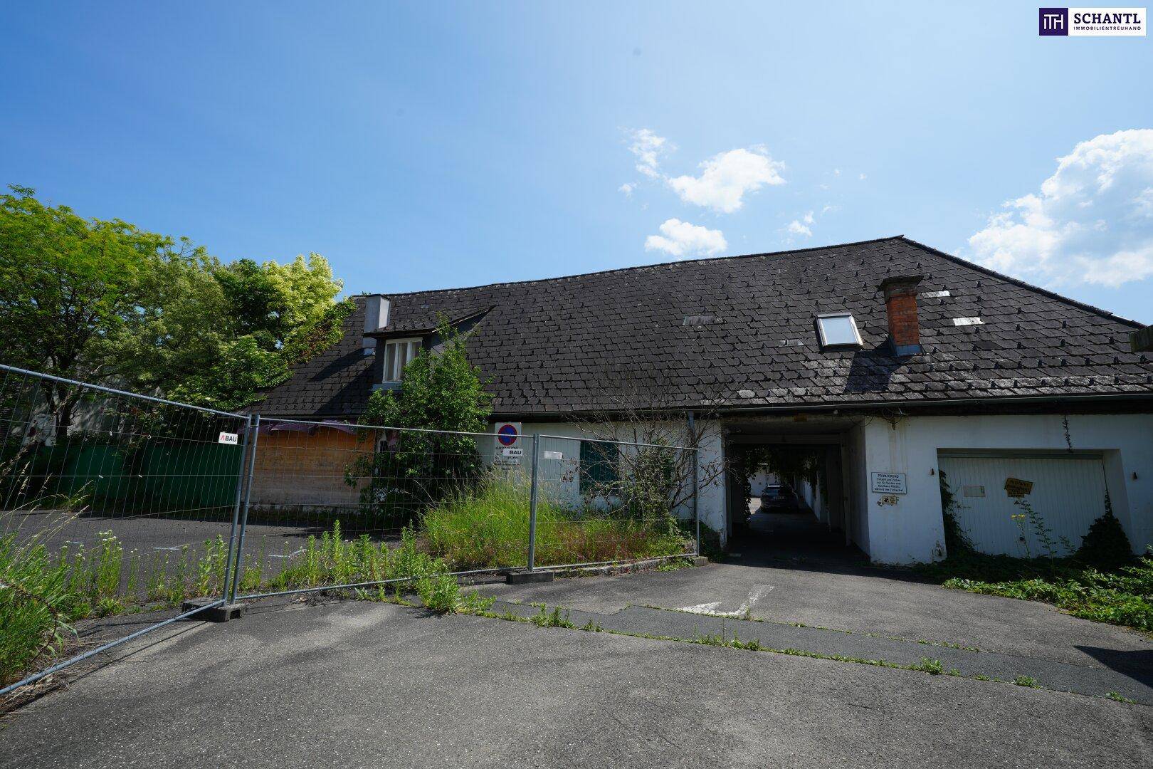 Altes Gebäude mit Dachgauben und einem Durchgang, umgeben von einem Bauzaun und Vegetation, wartet auf Sanierung.