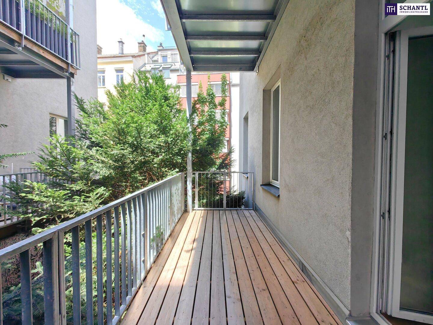 Heller Balkon mit Holzfußboden, Metallgeländer und Blick ins Grüne sowie auf umliegende Gebäude.