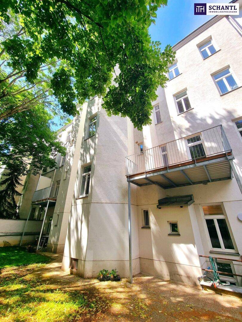 Helle Fassade eines Altbaus mit mehreren Balkonen und umgebendem Grünbereich unter blauem Himmel.