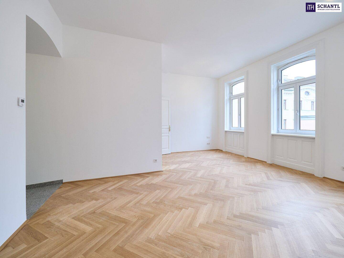 Geräumiger, heller Wohnraum mit klassischem Fischgrätparkett und zwei großen Fenstern.